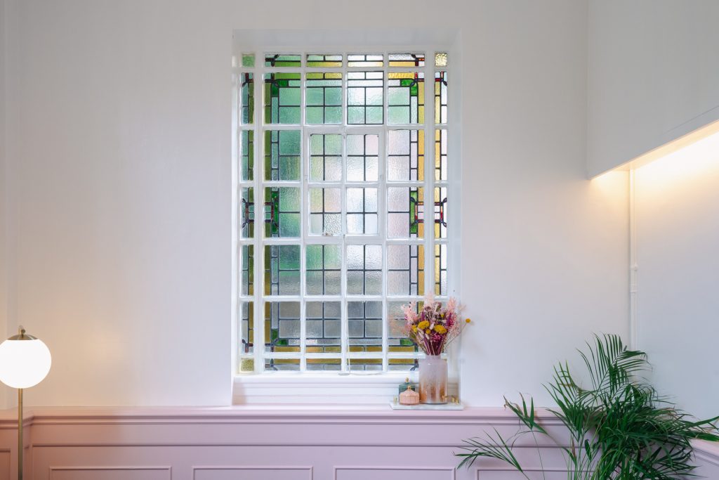 The stained glass window in The Canonbury Suite, as viewed from the couple's perspective, at Islington Town Hall, with pink dried flowers in a vase on the windowledge and a palm plant and a tall lamp on the floor in front of it