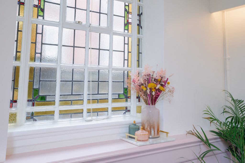 Detail of the stained glass window in The Canonbury Suite at Islington Town Hall, with pink dried flowers in a vase on the windowledge and a palm plant on the floor in front of it