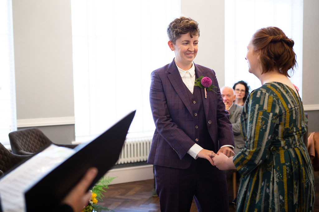 Two colourfully dressed brides hold hands and smile at one another while the registrar conducts their ceremony in Room 99 at Islington Town Hall