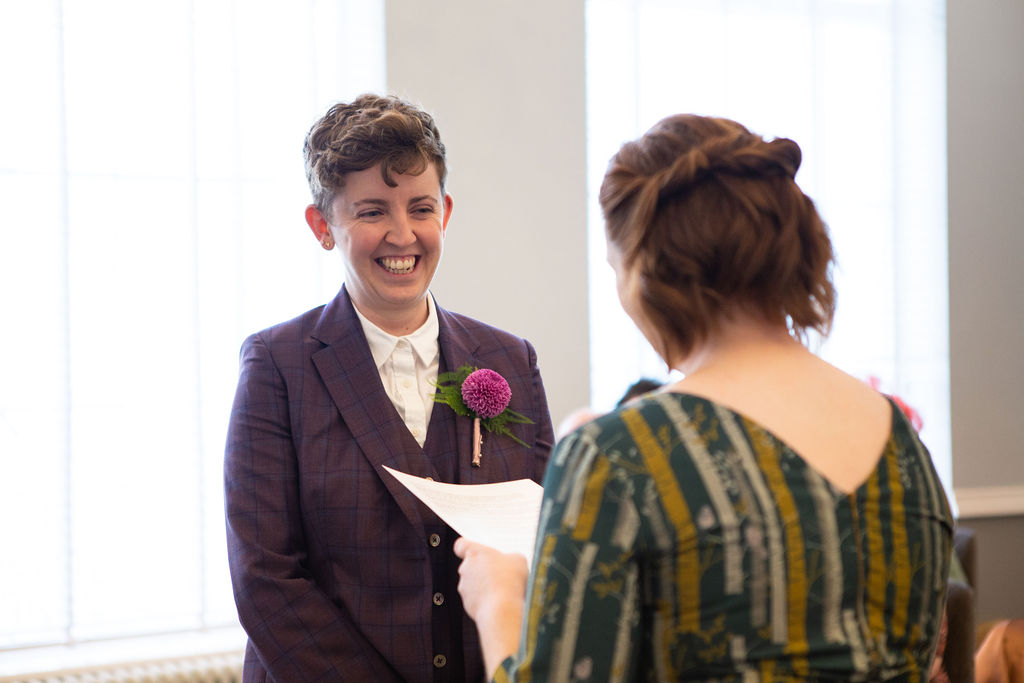 A bride in a purple suit smiles at her wife-to-be as she hears her personal vows for the first time in Room 99 at Islington Town Hall