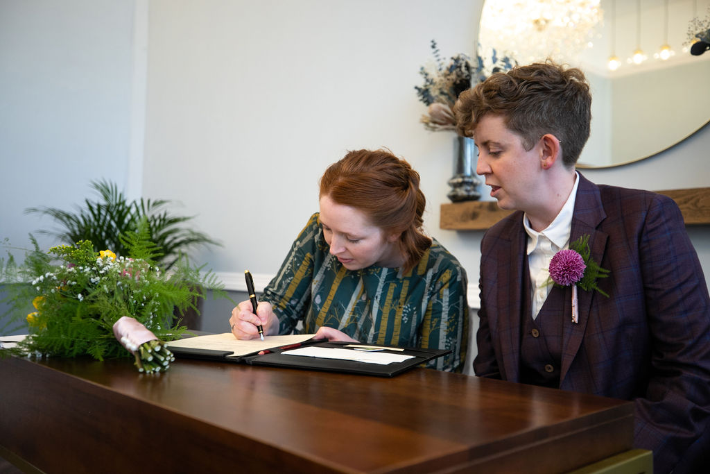 Two colourfully dressed brides sign the paperwork in Room 99 at Islington Town Hall