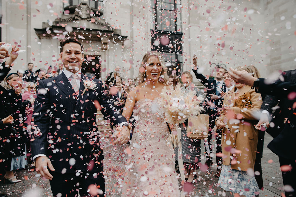 A groom in a dark suit and a bride in a pink dress leave the Town Hall in a shower of pink and white confetti
